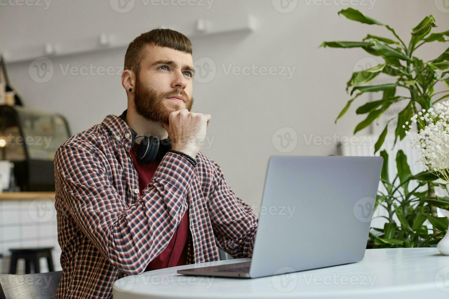 jovem atraente gengibre barbudo homem senta às uma mesa dentro uma cafeteria e trabalho em uma computador portátil, vestindo dentro básico roupas, pensativamente olhando ausente. foto