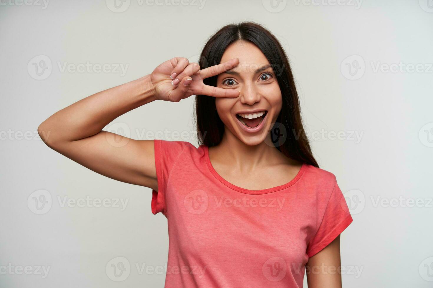 alegre jovem adorável Castanho cabelos mulher com casual Penteado levantando vitória gesto para dela face e sorridente alegremente para Câmera, vestindo Rosa camiseta enquanto posando sobre branco fundo foto