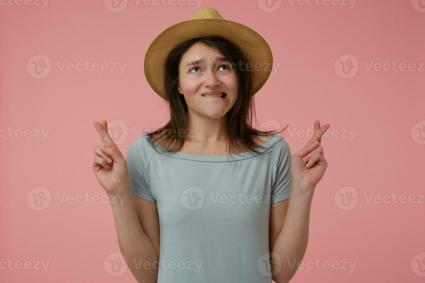 retrato do atraente, adulto menina com grandes morena cabelo. vestindo azulado camiseta e chapéu. olhando acima, morder lábio e fazer uma desejar, dedos cruzado. ficar de pé isolado sobre pastel Rosa fundo foto