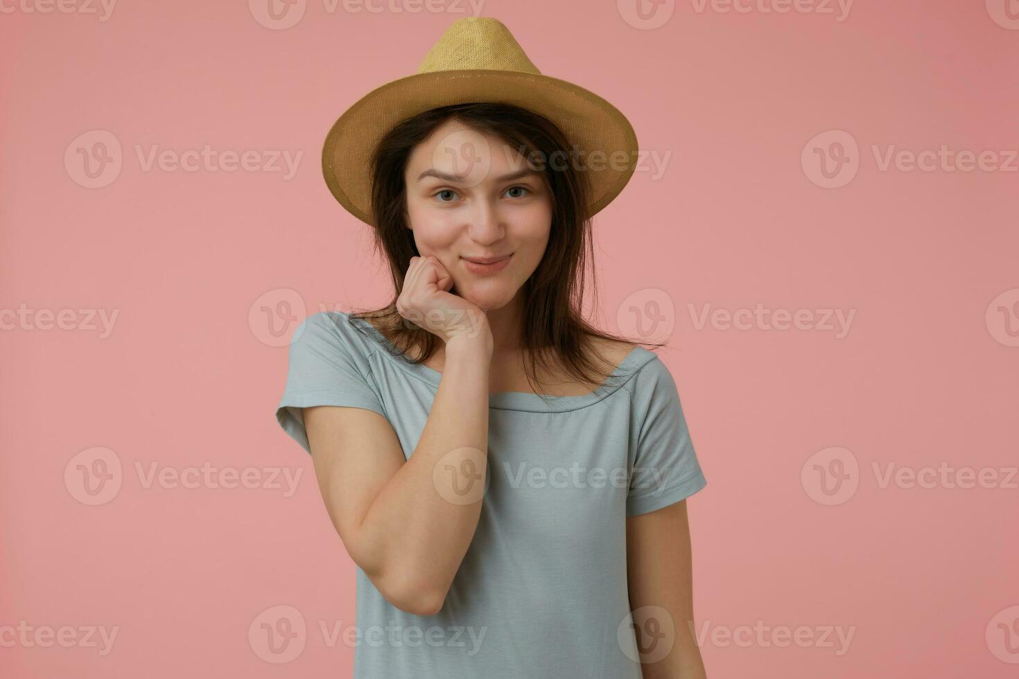 agradável olhando mulher, envergonhado menina com grandes morena cabelo. vestindo azulado camiseta e chapéu. segurando mão debaixo dela queixo e parece provocante. assistindo às a Câmera isolado sobre pastel Rosa fundo foto