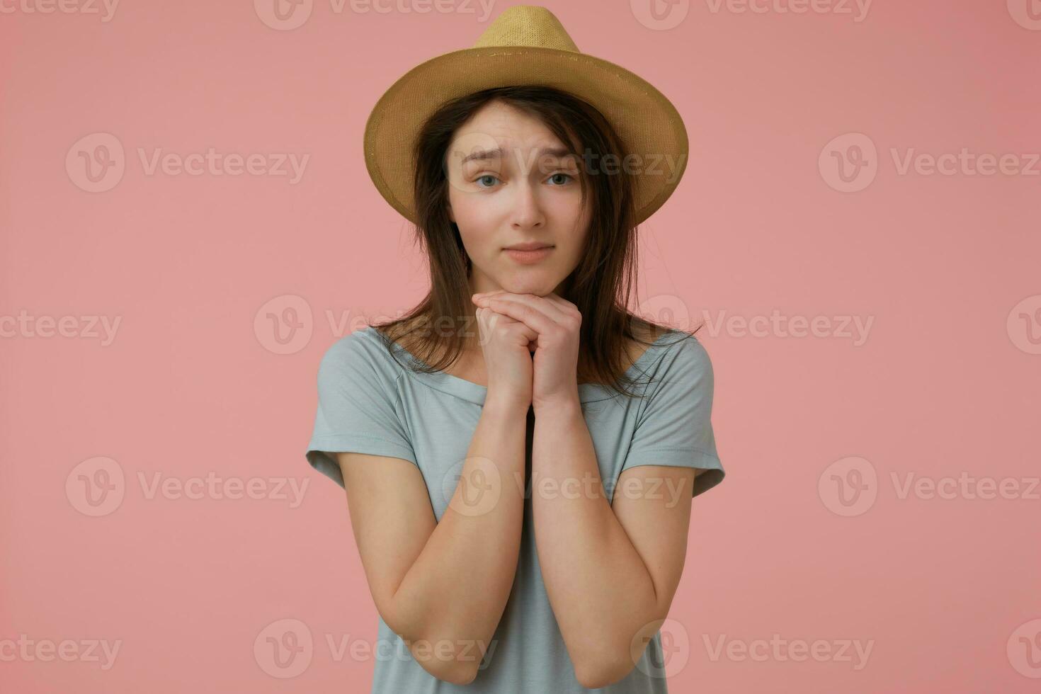 agradável olhando mulher, lindo menina com grandes morena cabelo. vestindo azulado camiseta e chapéu. Veja às você e implorar para algo. assistindo às a Câmera isolado sobre pastel Rosa fundo foto