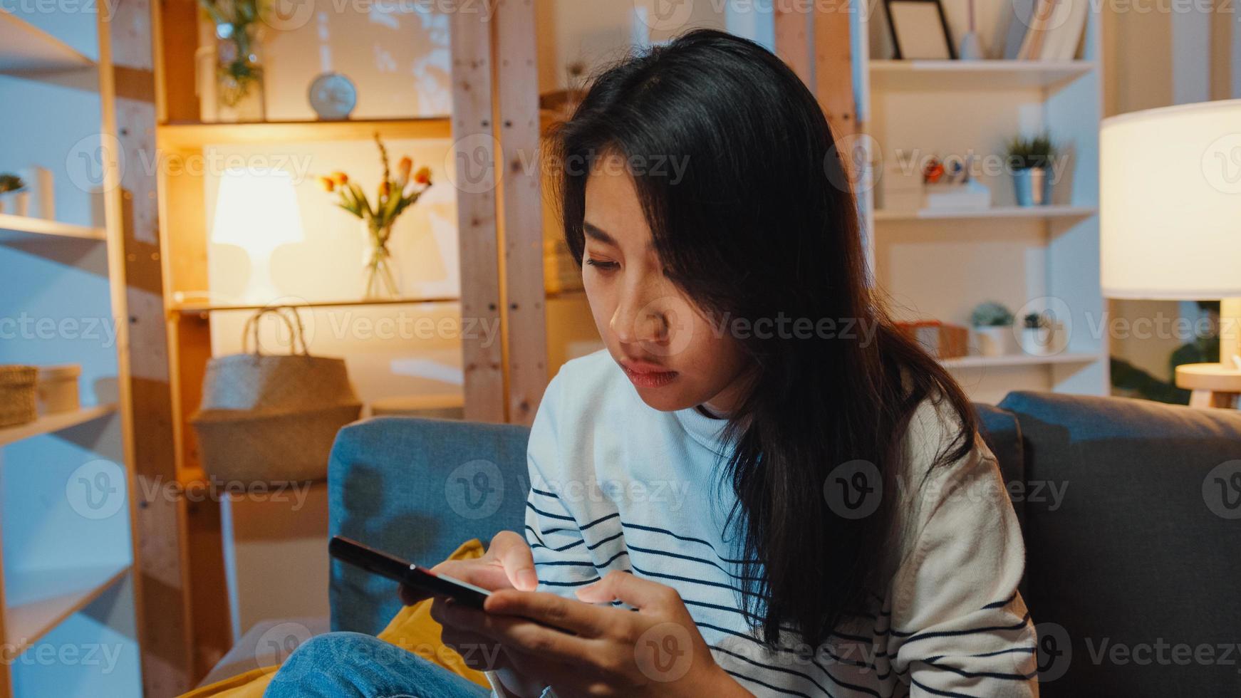 senhora asiática, pensativa, segurando o telefone, sentindo-se triste à espera de uma ligação, sente-se no sofá na sala de estar à noite em casa, sinta-se solitária, triste adolescente deprimido passar um tempo sozinho, distância social, quarentena de coronavírus. foto