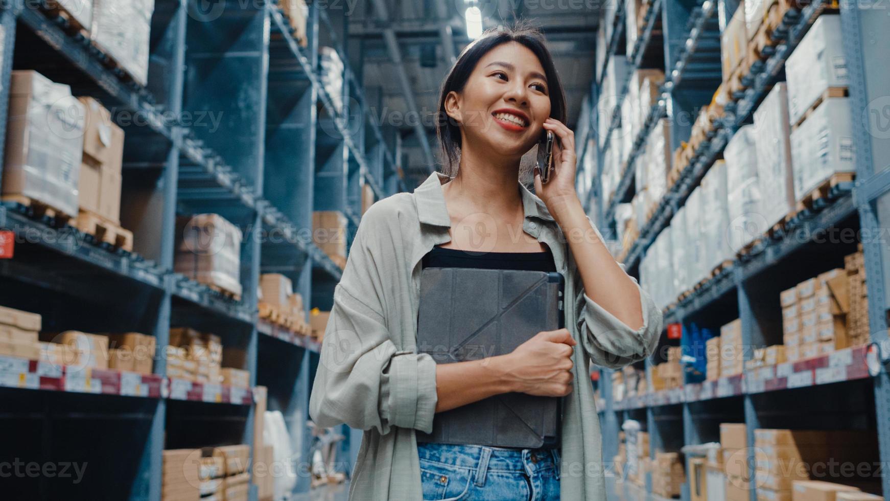 vendedor on-line de jovem empresária da ásia falando em confirmação de pedidos do cliente no telefone, segurando o tablet digital em pé no centro comercial de varejo. distribuição, logística, embalagens prontas para embarque. foto