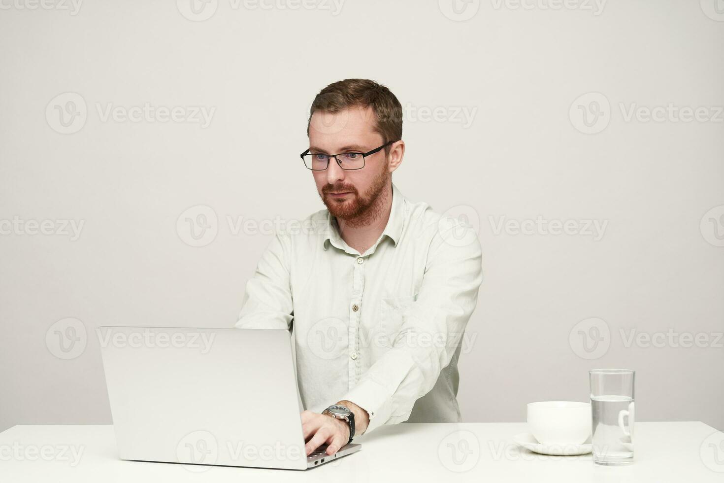 concentrado jovem barba por fazer cabelo curto masculino dentro óculos digitando texto com dele computador portátil e olhando atentamente em tela, sentado sobre branco fundo foto