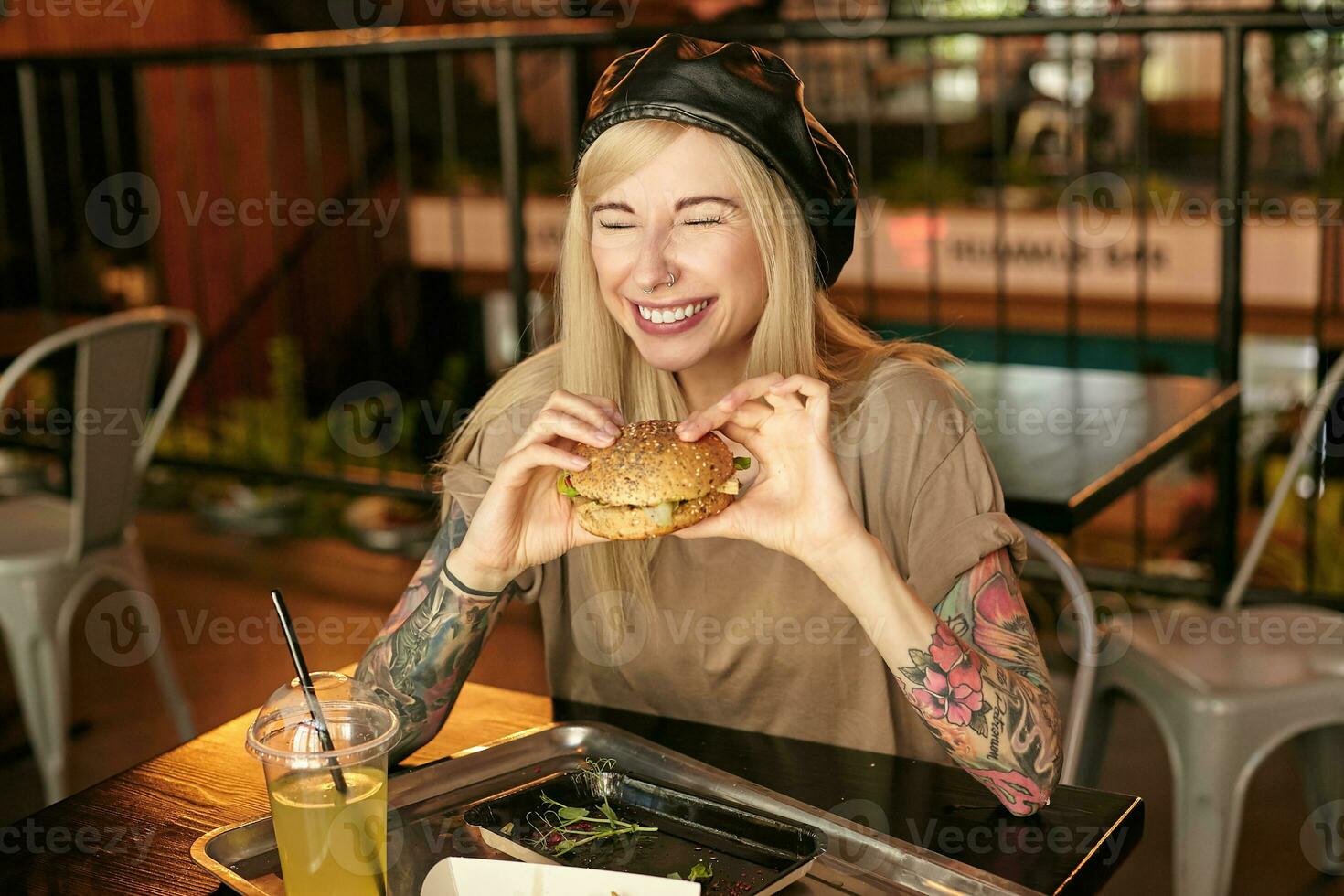 feliz jovem Loiras mulher com tatuagens tendo almoço dentro cidade cafeteria, sentado às mesa com hamburguer dentro mãos e rindo alegremente com fechadas olhos, vestindo na moda roupas foto