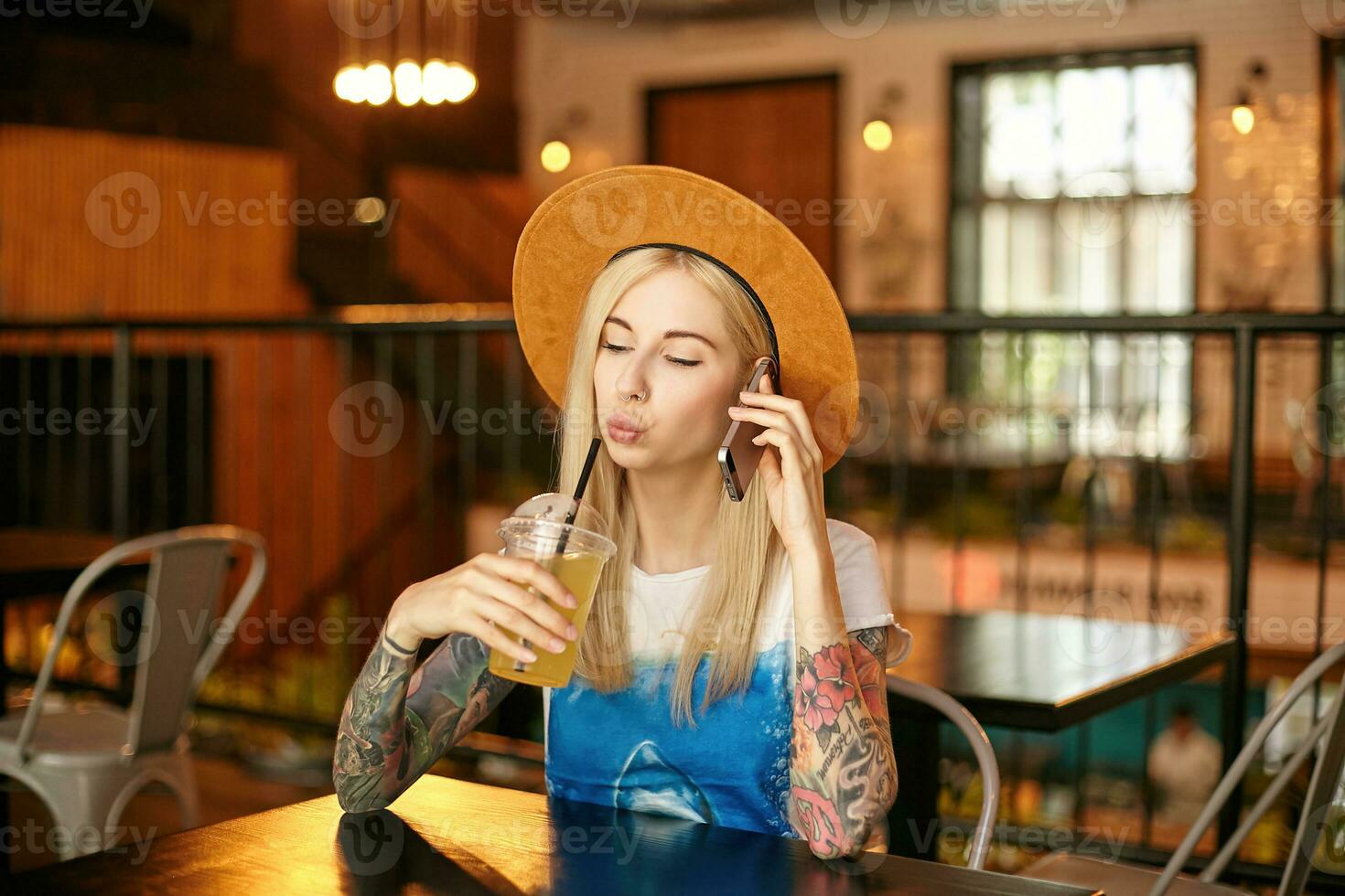 horizontal tiro do atraente tatuado jovem mulher dentro casual roupas sentado às mesa dentro cidade cafeteria e bebendo fresco suco, guardando Móvel telefone dentro mão e fazer ligar foto