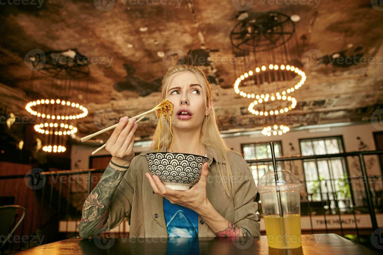 retrato do duvidando tatuado Loiras fêmea com casual Penteado tentando primeiro ásia comida, segurando carâmico tigela com Macarrão e olhando a parte, de lado com confuso face foto