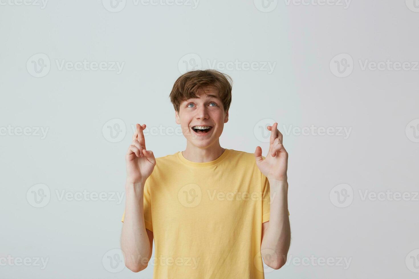 retrato do feliz atraente jovem homem com suspensórios em dentes dentro amarelo t camisa mantém dedos cruzado e fazer uma desejo isolado sobre branco fundo foto