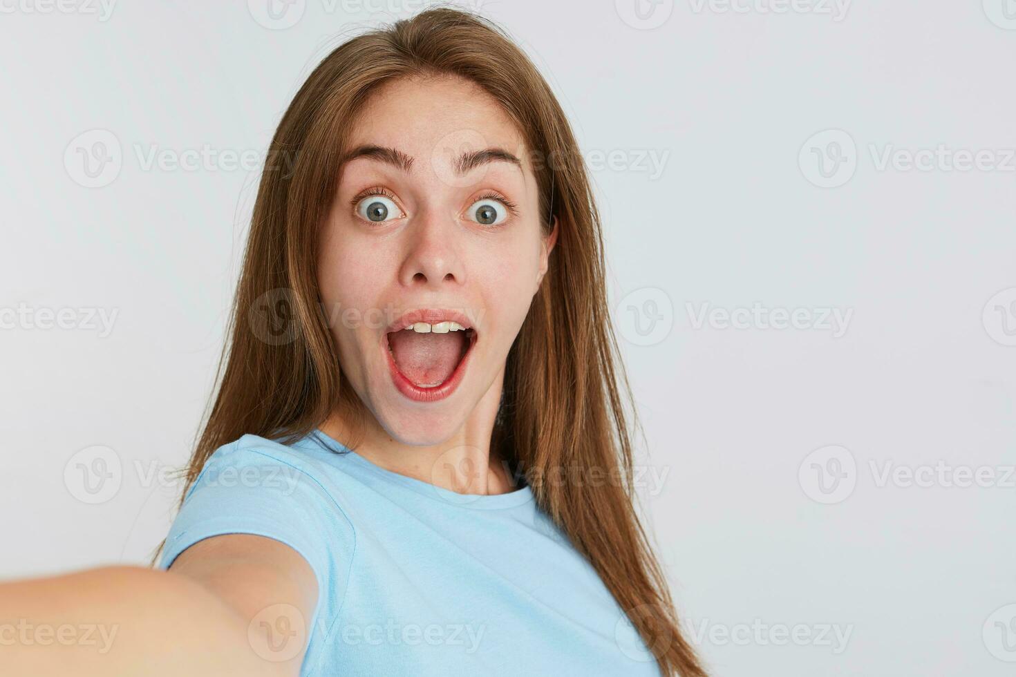 fechar-se do feliz surpreso jovem mulher com aberto boca desgasta azul t camisa parece espantado e fazer selfie isolado sobre branco fundo foto