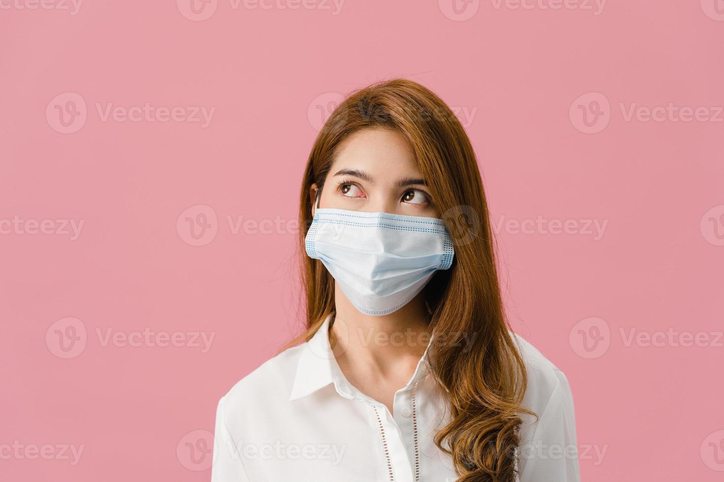 jovem asiática usando máscara médica com vestido de pano casual e olhando para o espaço em branco isolado no fundo rosa. auto-isolamento, distanciamento social, quarentena para prevenção do vírus corona foto