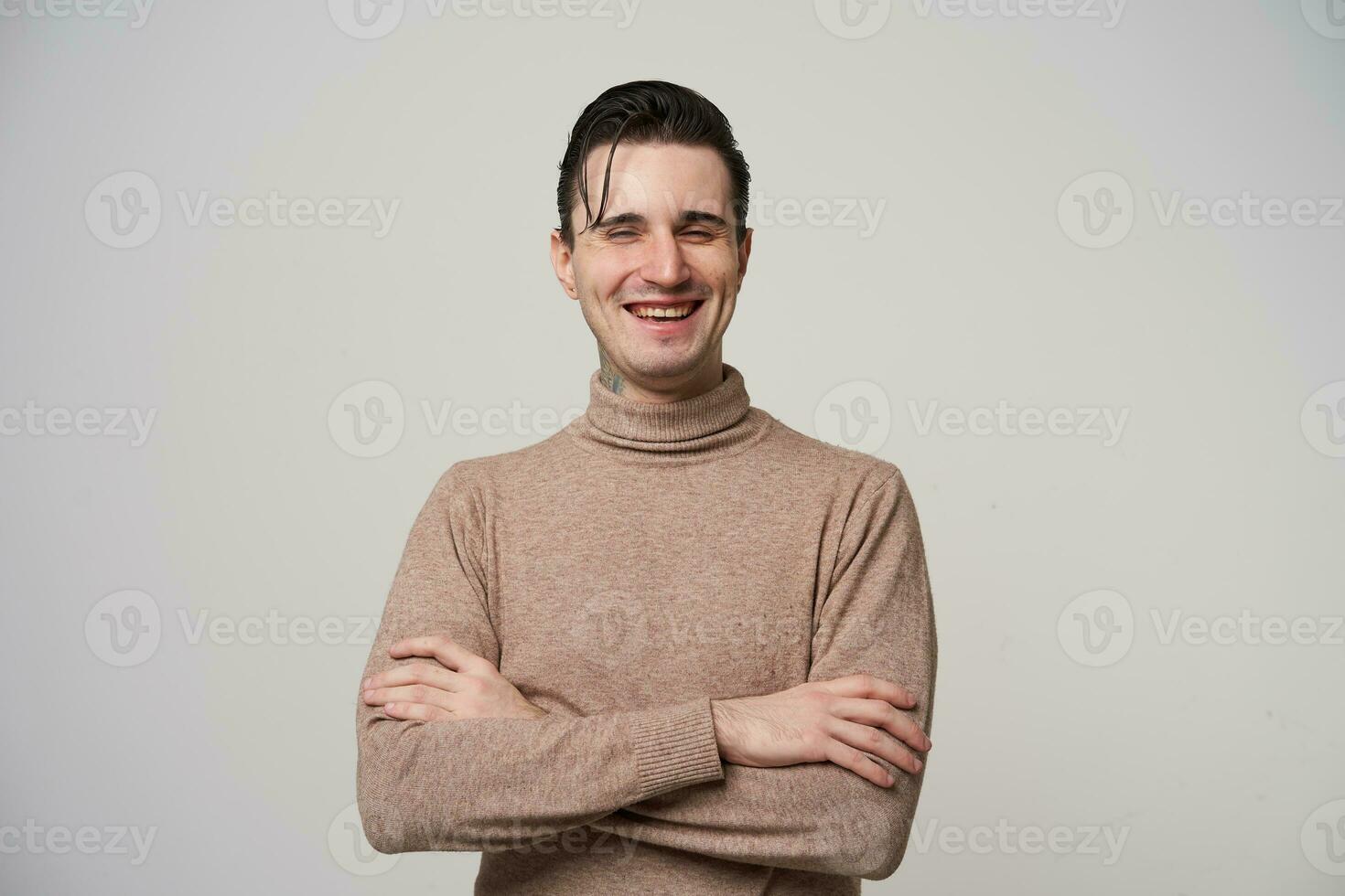 alegre lindo jovem Sombrio cabelos homem com guardada mãos rindo alegremente com fechadas olhos enquanto posando sobre branco fundo, vestindo bege pescoço enrolado suéter foto