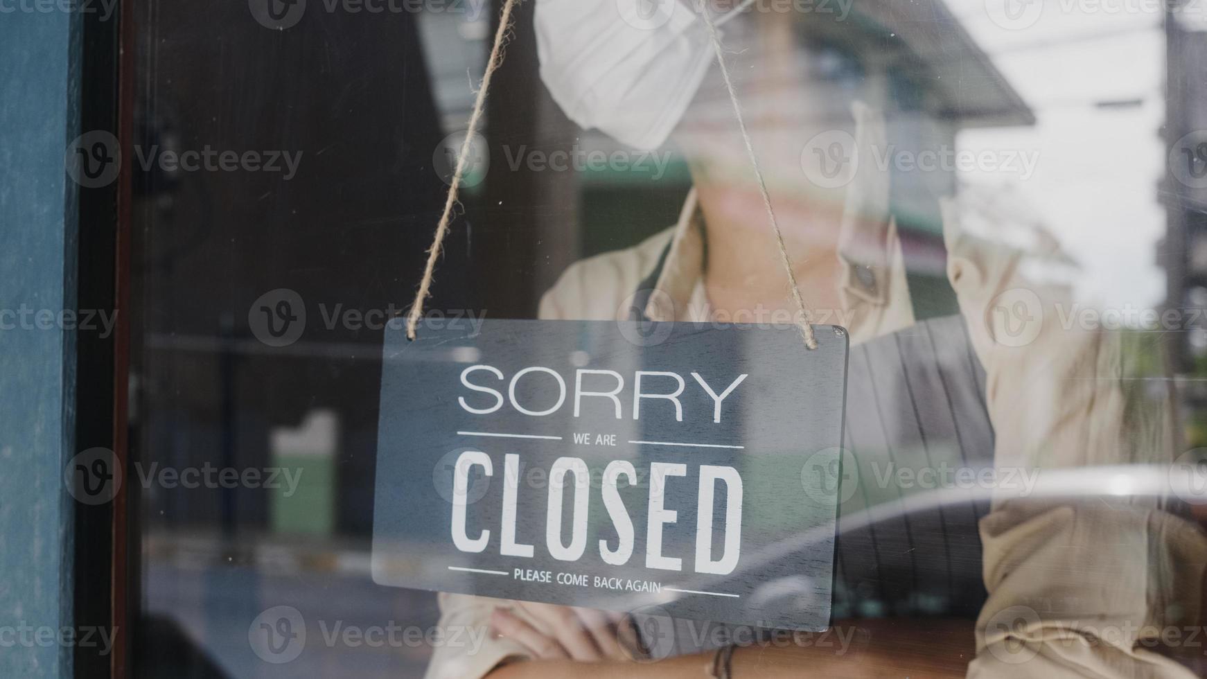 jovem asiática usa máscara facial virando uma placa de aberta para fechada na porta de vidro do café após a quarentena de bloqueio de coronavírus. proprietário de uma pequena empresa, comida e bebida, conceito de crise financeira empresarial foto