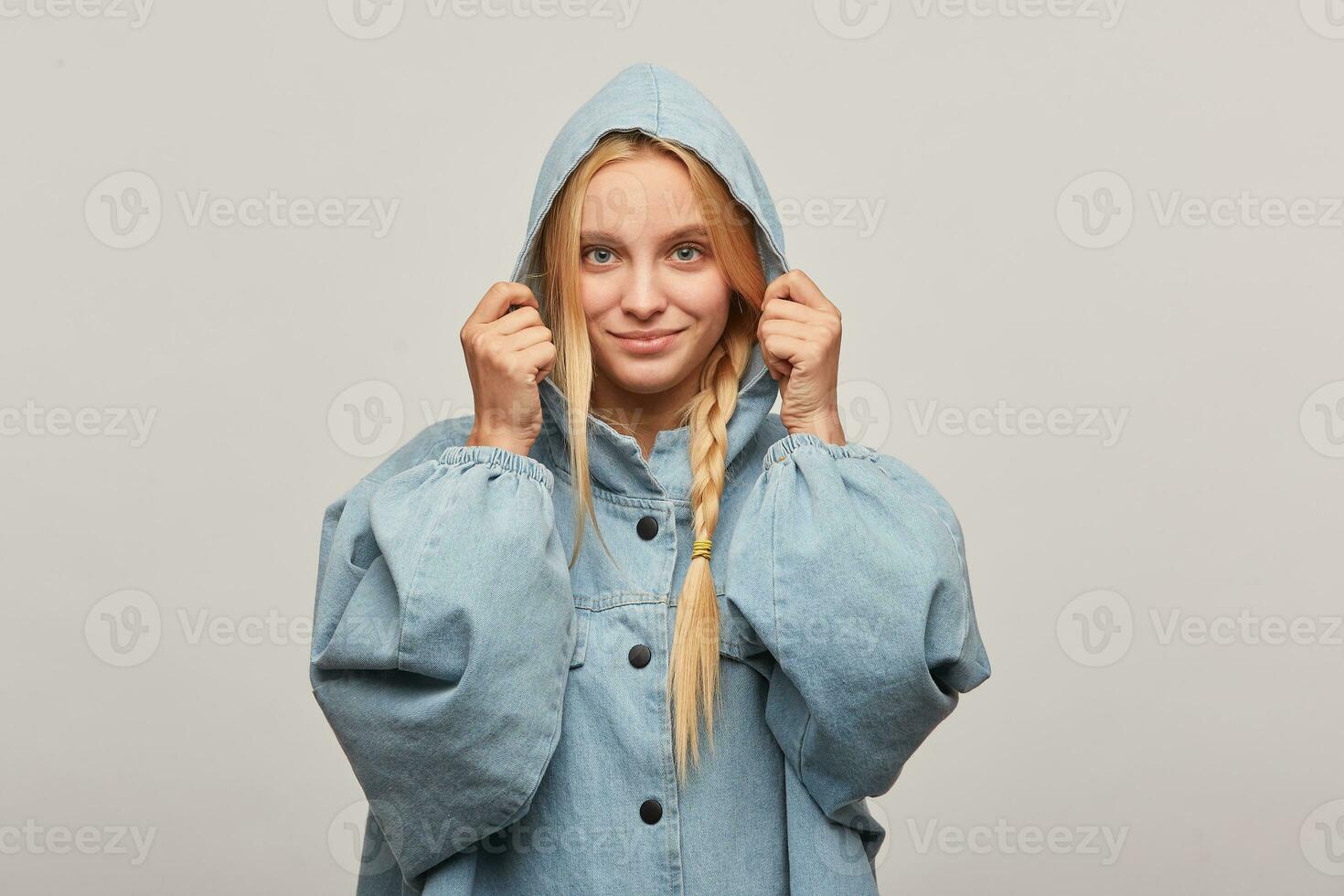 estúdio tiro do adorável lindo Loiras mulher com trança, mãos manter capuz, sente feliz ternamente sorrisos, desgasta grande demais jeans jeans casaco sentindo-me aquecido não receoso do frio clima, sobre branco fundo foto