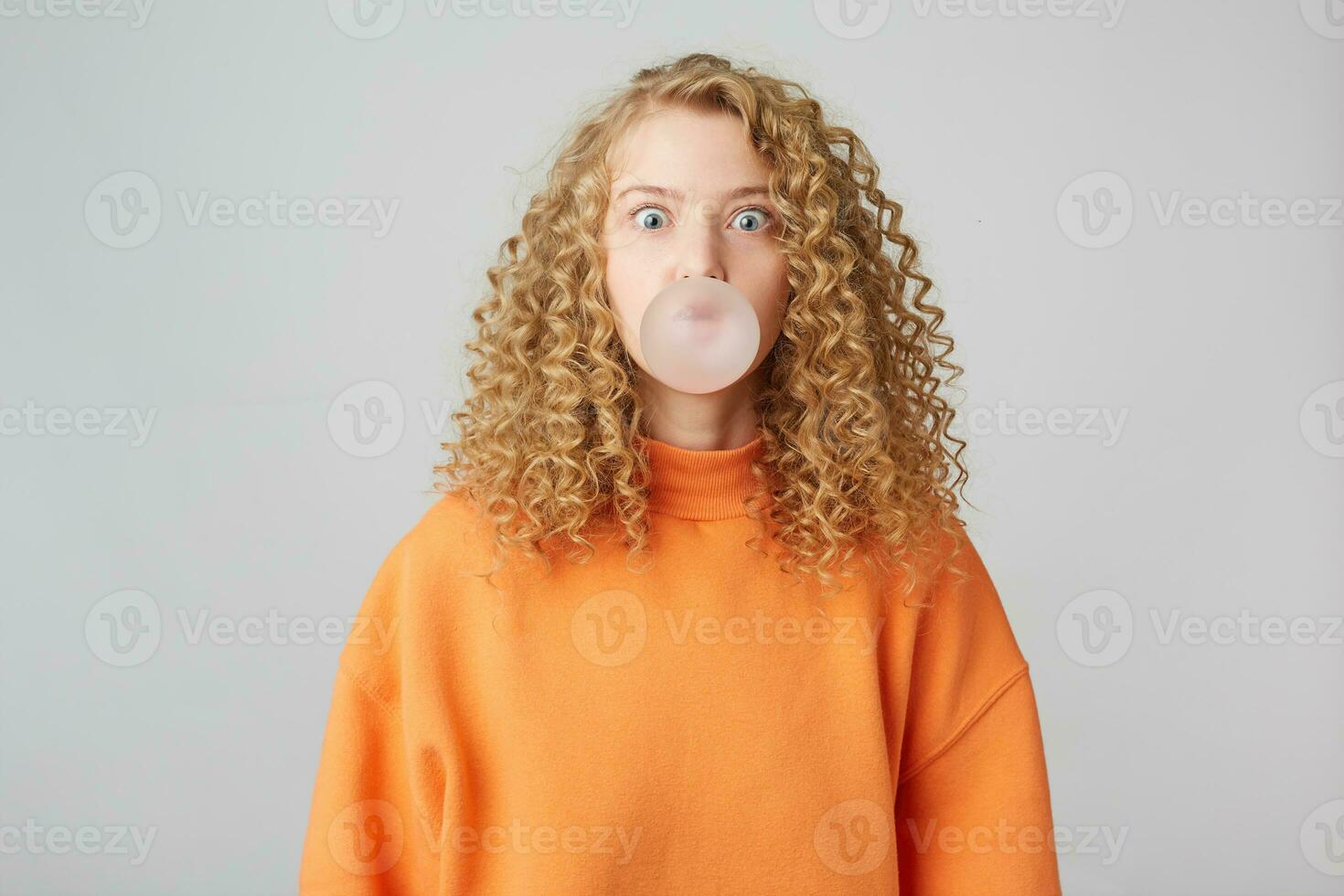 encarando dentro Câmera Loiras sopro bolha com mastigar Chiclete, desgasta dentro caloroso brilhante laranja grande demais suéter em pé isolado sobre branco fundo foto