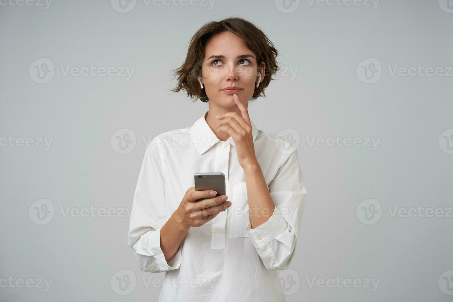 pensativo jovem bonita fêmea com casual Penteado olhando para cima e guardando indicador em dela lábio inferior, posando sobre branco fundo com Smartphone dentro dela mão foto