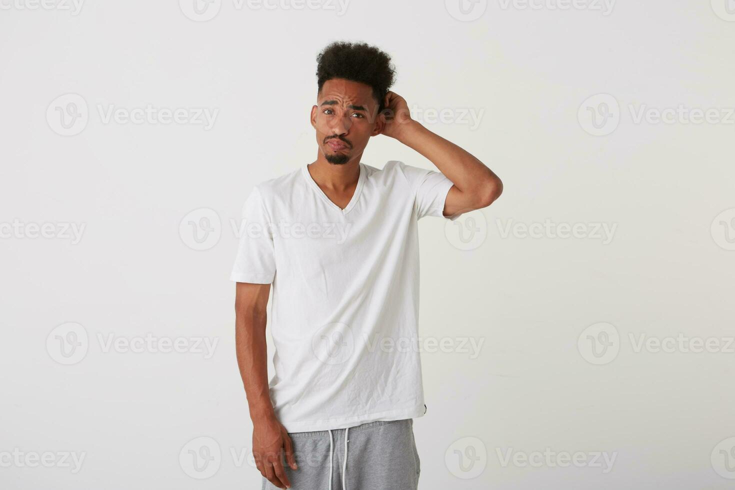 estúdio foto do jovem bonita Sombrio esfolado morena homem vestido dentro casual roupas fazendo careta dele face e coçar cabeça enquanto em pé contra branco fundo