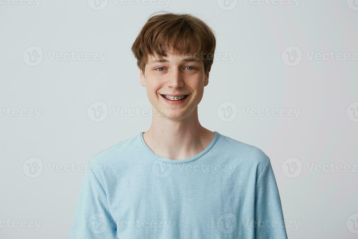 adolescente com curto cabelo e suspensórios em dentes parece Câmera, desgasta azul camiseta, sente feliz alegre, sorridente isolado sobre branco fundo foto