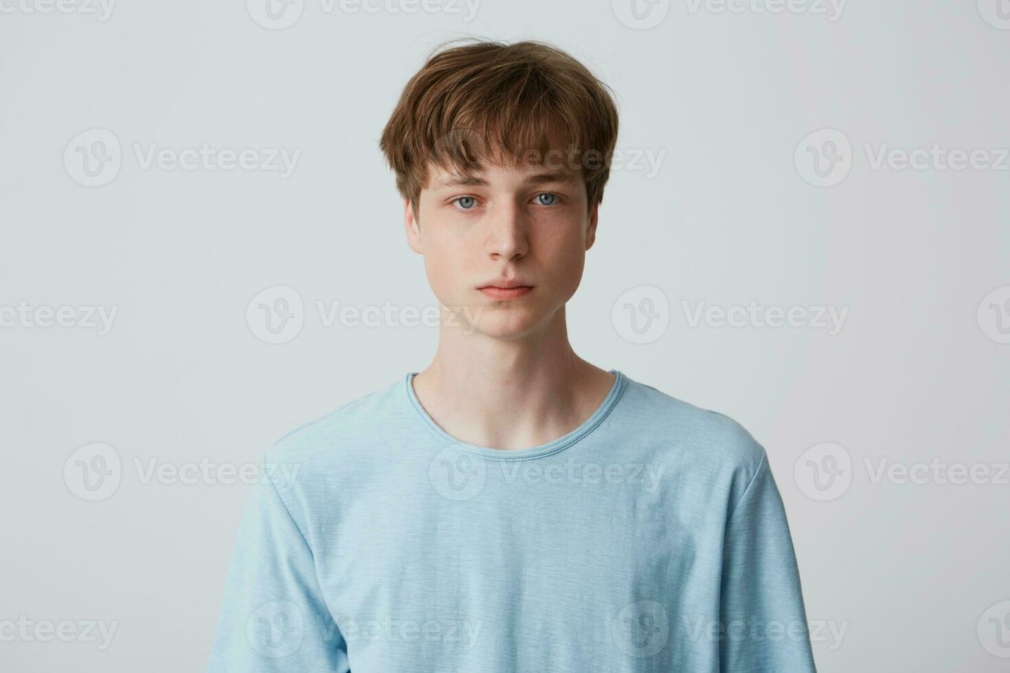 fechar acima face do uma jovem homem sem emoções. lindo sem emoção cara dentro uma azul camiseta olhando para a Câmera, isolado sobre branco fundo foto