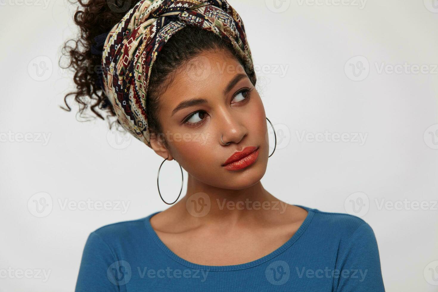 interior tiro do jovem bonita Sombrio cabelos encaracolado senhora inclinação dela cabeça enquanto olhando maravilhosamente aparte, vestido dentro azul blusa e colori arco de cabelo enquanto posando sobre branco fundo foto