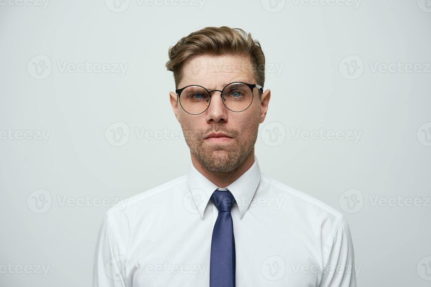 retrato do jovem homem de negocios olhando desconfiadamente, sente incerto e dúvida, com à moda corte de cabelo, copos, barba por fazer, vestido dentro perfeitamente passado a ferro branco camisa e azul amarrar, acabar branco fundo foto