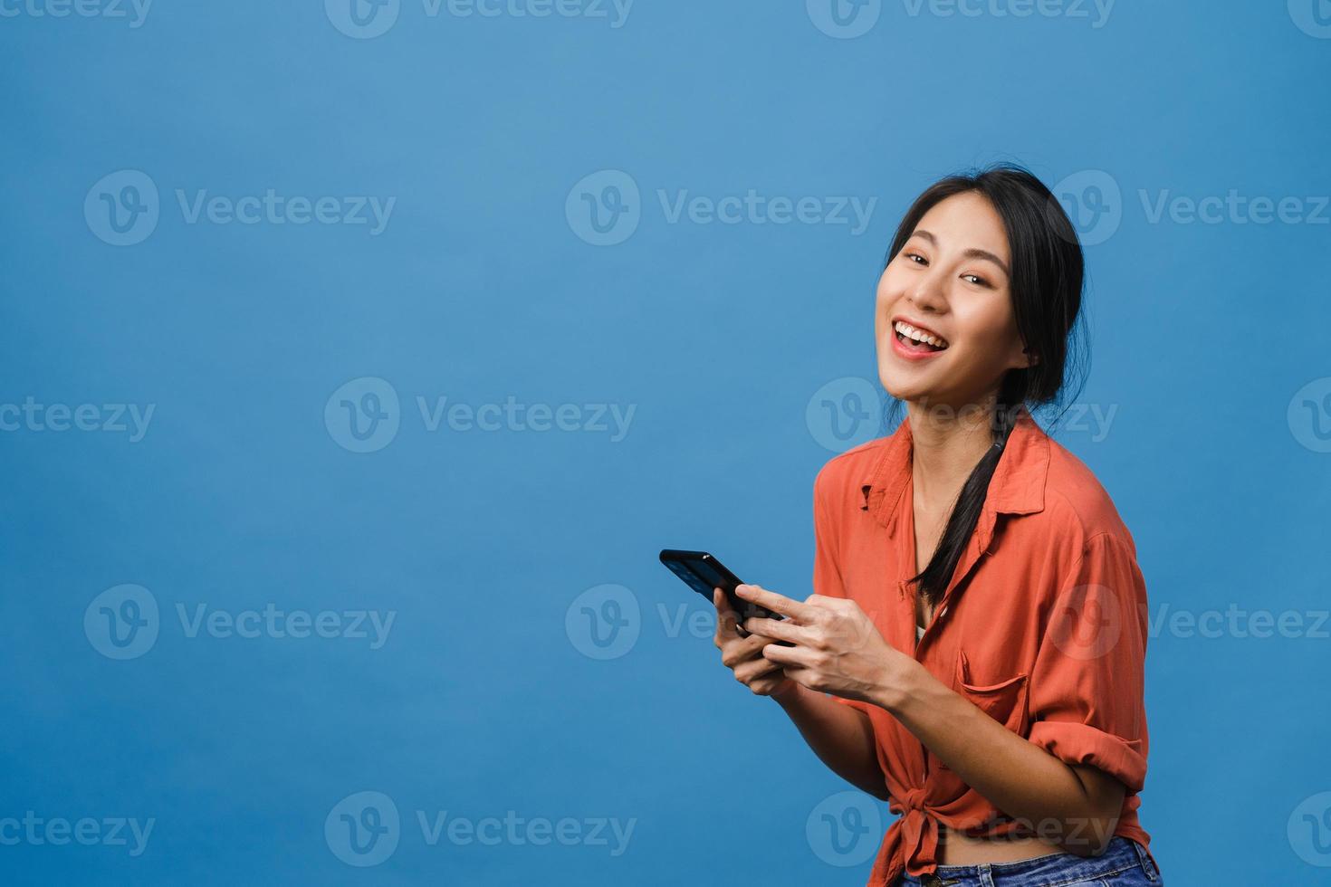 surpreendeu a jovem asiática usando telefone celular com expressão positiva, sorria amplamente, vestida com roupas casuais e olhando para a câmera sobre fundo azul. feliz adorável feliz mulher alegra sucesso. foto