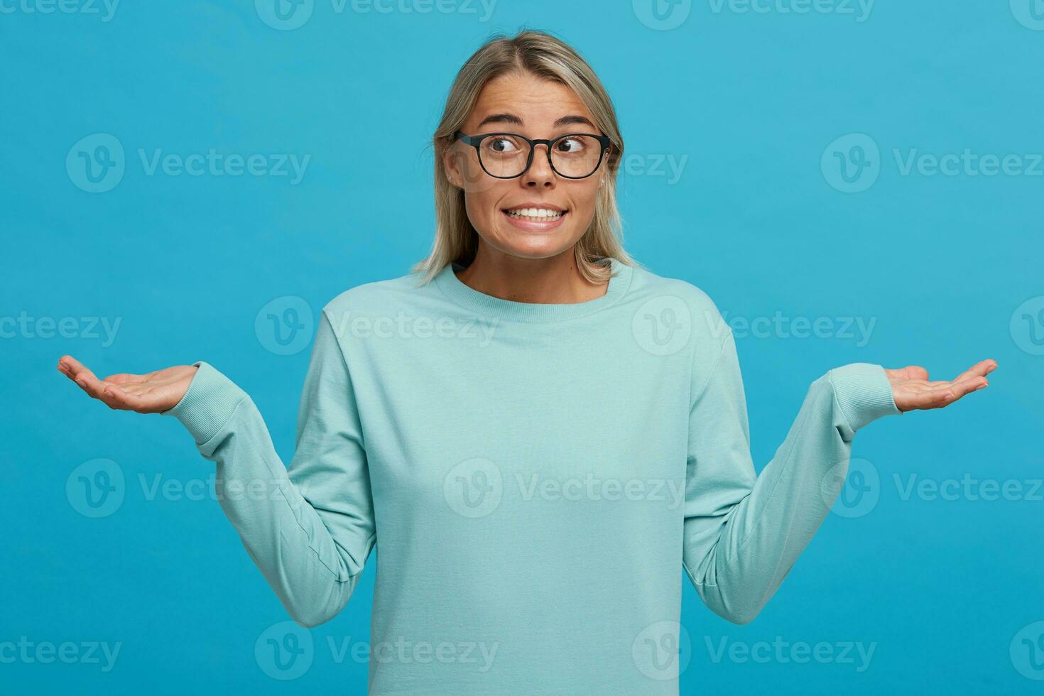 confuso jovem menina encolhendo os ombros ombros, tendo culpado olhar, sentindo-me Desculpe para fazendo alguma coisa errado e fazer Terrível erro, vestido dentro luz azul grandes manga camiseta, sobre azul fundo foto