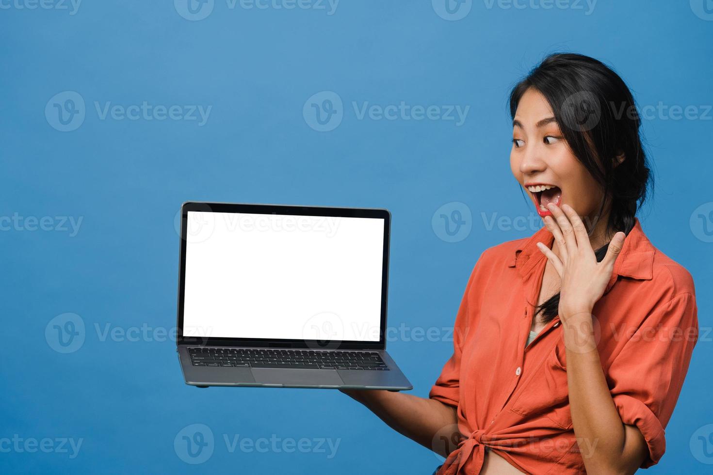 jovem asiática mostra a tela do laptop vazia com expressão positiva, sorri amplamente, vestida com roupas casuais, sentindo felicidade isolada sobre fundo azul. computador com tela branca na mão feminina. foto