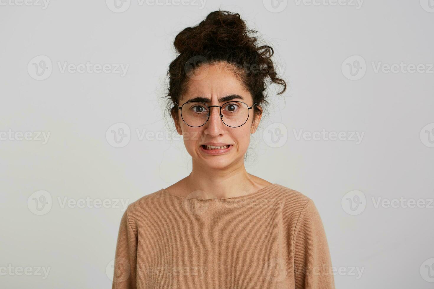 retrato do envergonhado preocupado jovem mulher com pão do Sombrio encaracolado cabelo desgasta bege suéter e óculos sente confuso e preocupado isolado sobre branco fundo foto