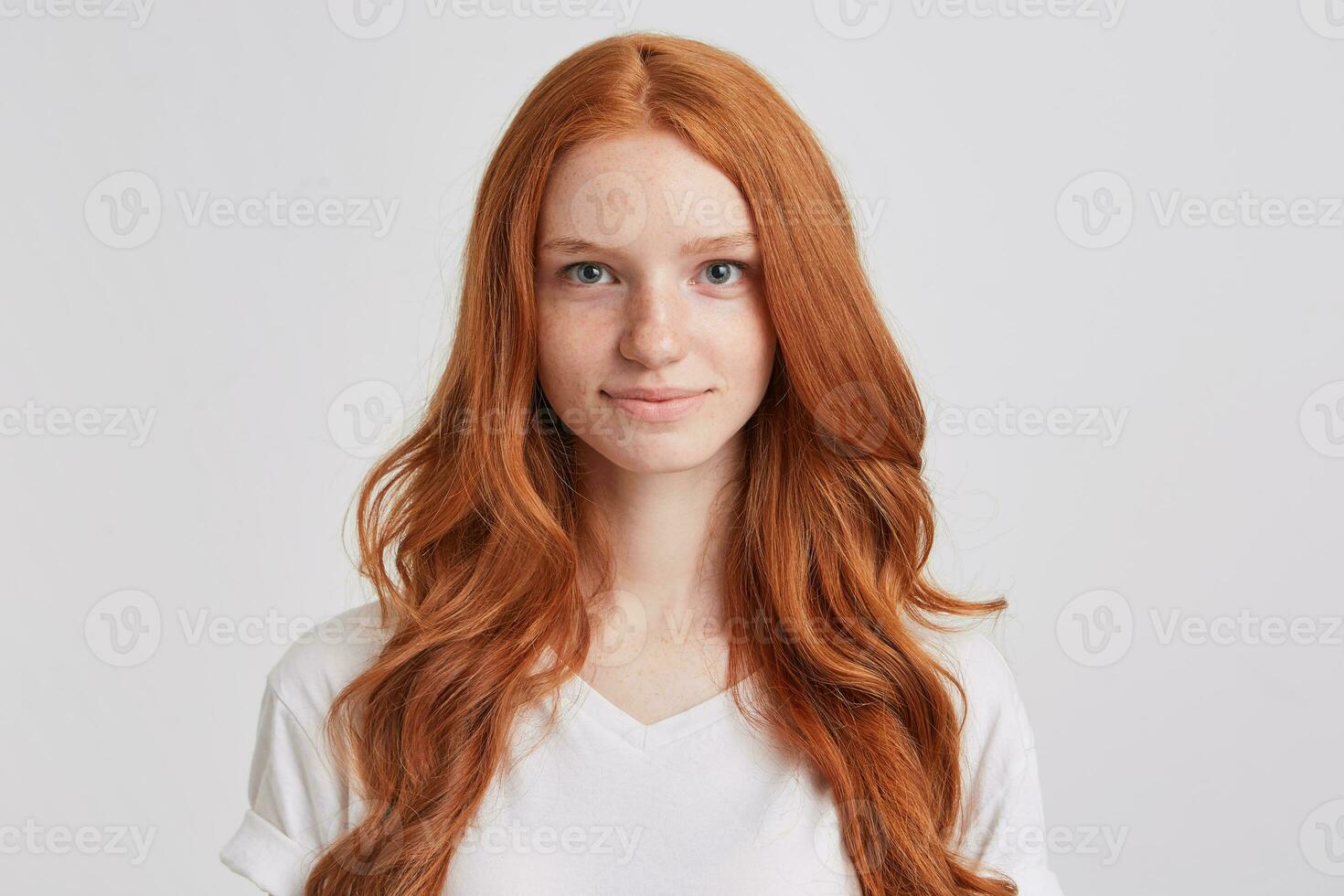 retrato do sorridente atraente ruiva jovem mulher com grandes ondulado cabelo e sardas desgasta t camisa sente feliz e satisfeito isolado sobre branco fundo parece diretamente dentro Câmera foto