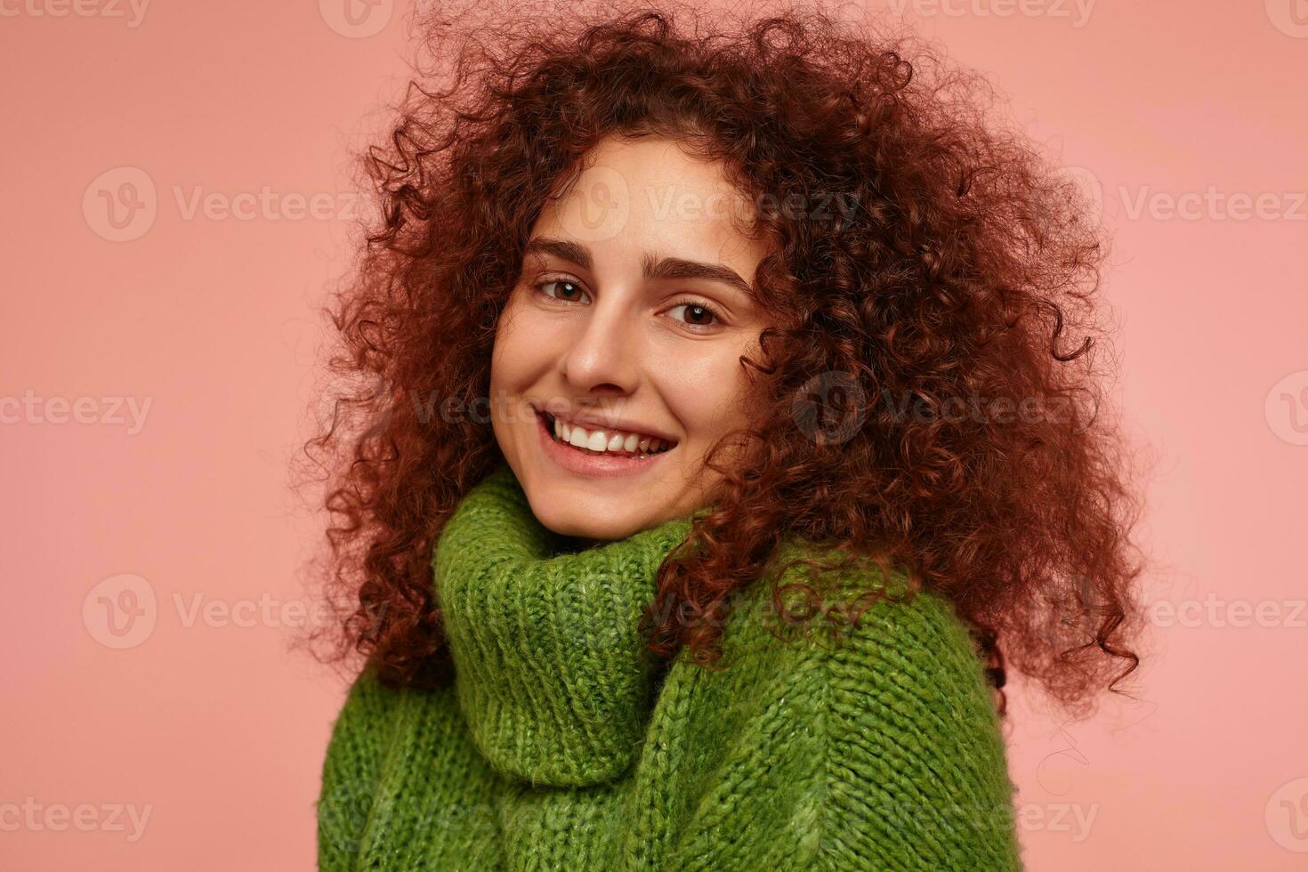 retrato do atraente, adulto menina com gengibre encaracolado cabelo. vestindo verde gola alta suéter e sorridente. assistindo provocante às a Câmera isolado, fechar-se sobre pastel Rosa fundo foto