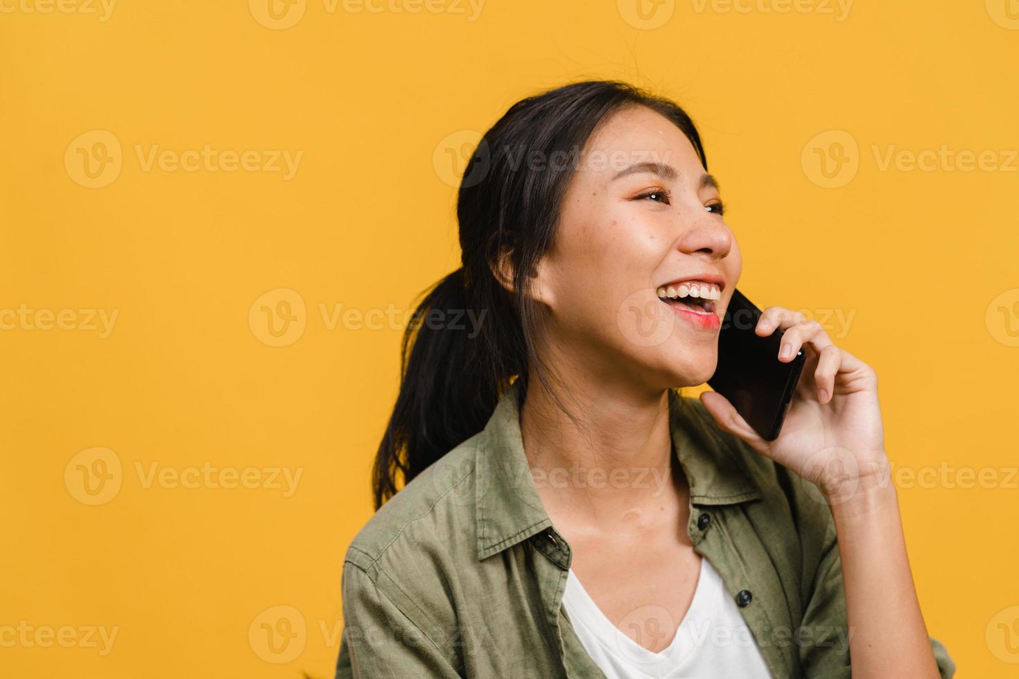 jovem asiática fala por telefone com uma expressão positiva, sorri amplamente, vestida com roupas casuais, sentindo felicidade e fica isolado em um fundo amarelo. feliz adorável feliz mulher alegra sucesso. foto