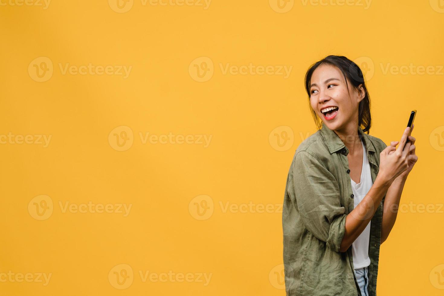 jovem asiática usando telefone com expressão positiva, sorri amplamente, vestida com roupas casuais, sentindo felicidade e carrinho isolado em fundo amarelo. feliz adorável feliz mulher alegra sucesso. foto