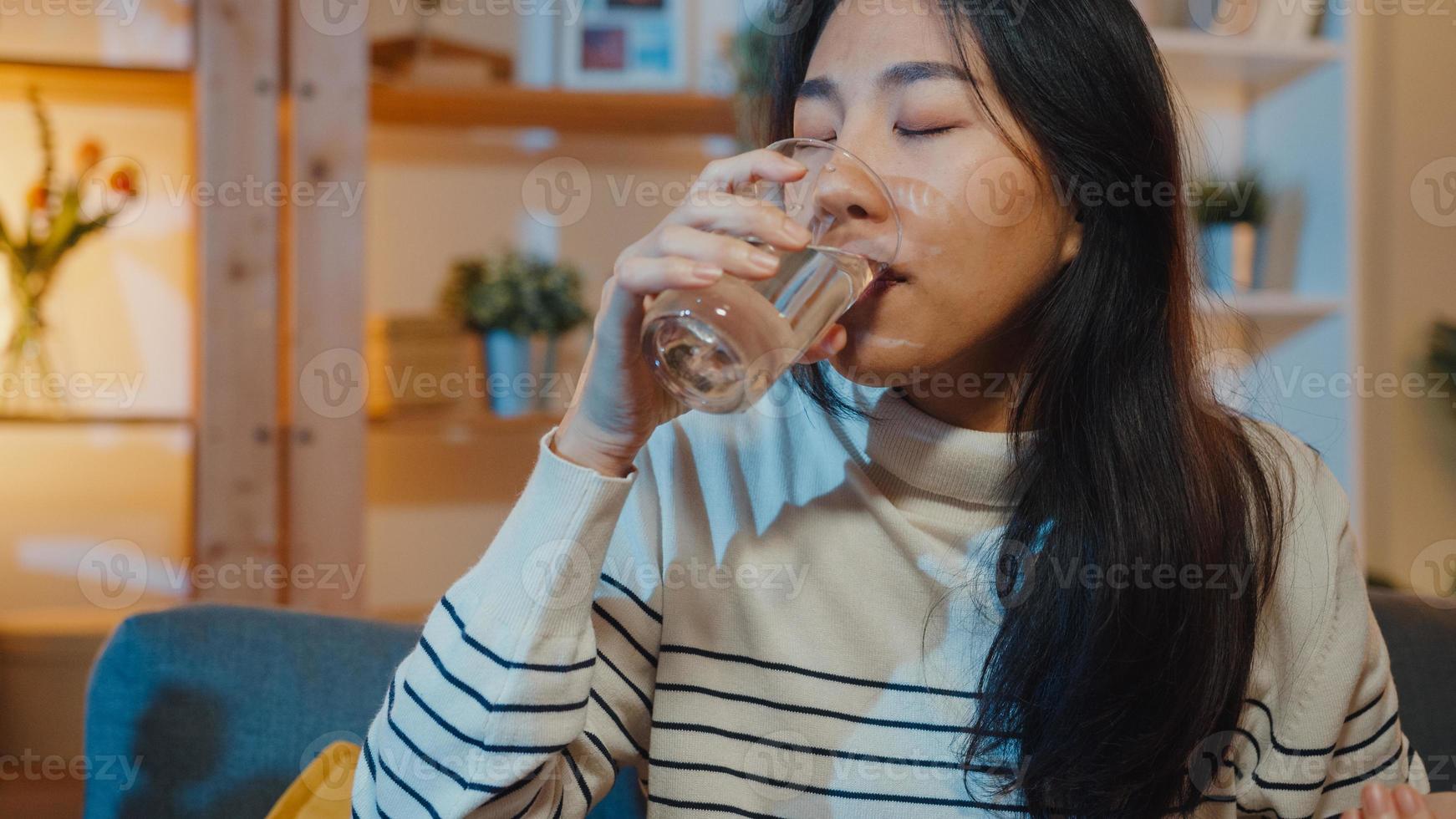 mulher asiática jovem doente segurando um copo de comprimido de água tomar remédio sentar no sofá em casa à noite. garota tomando remédio após ordem do médico, quarentena em casa, conceito de distanciamento social de coronavírus. foto