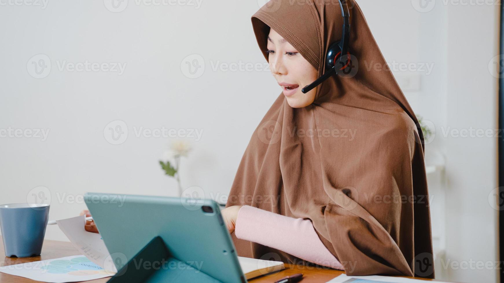 A senhora muçulmana asiática usa fone de ouvido usando tablet digital e fala com colegas sobre o relatório de venda em videoconferência enquanto trabalha em casa na cozinha. distanciamento social, quarentena para o vírus corona. foto