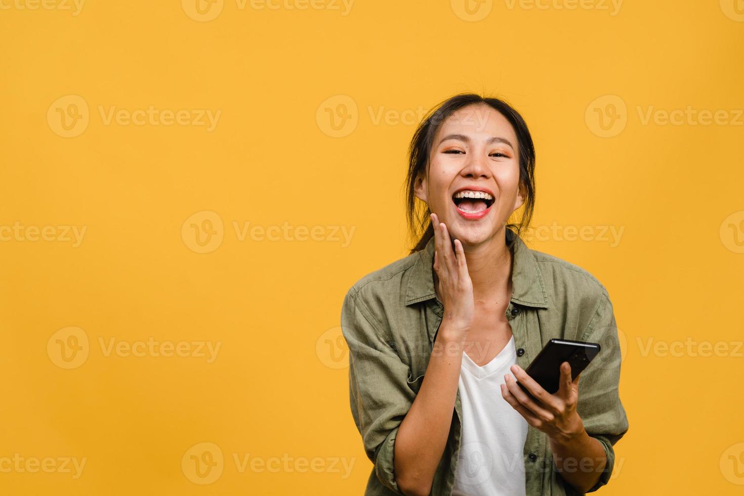 surpreendeu a jovem asiática usando telefone celular com expressão positiva, sorri amplamente, vestida com roupas casuais e carrinho isolado em fundo amarelo. feliz adorável feliz mulher alegra sucesso. foto