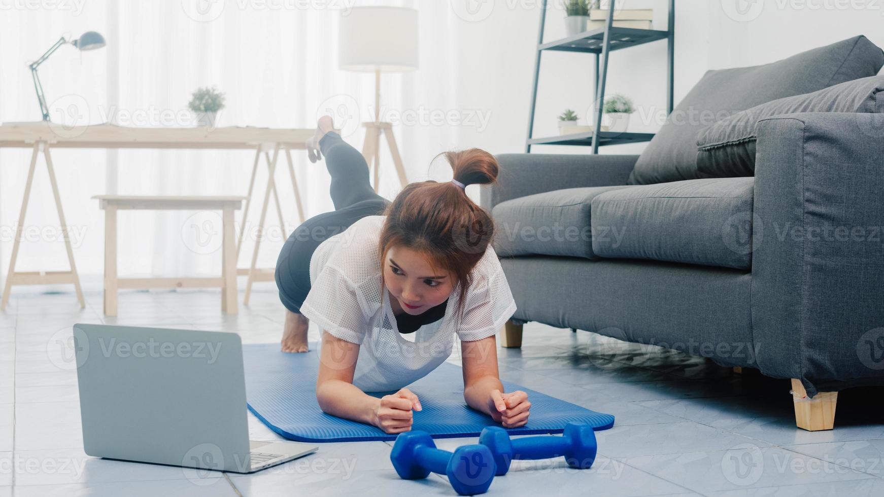 jovem senhora em exercícios de sportswear fazendo malhar pranchas com uma perna estendida e usando o laptop para assistir a um vídeo tutorial de ioga em casa. treino à distância com personal trainer, distância social. foto