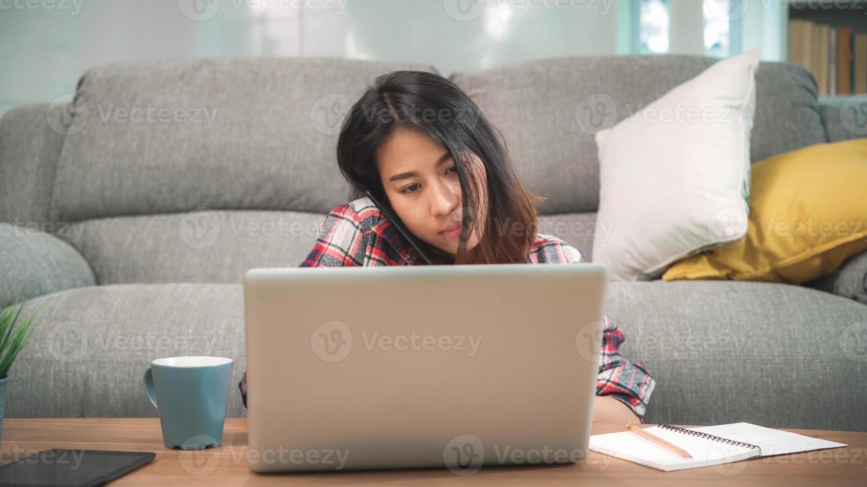 mulher asiática freelance trabalhando em casa, mulher de negócios trabalhando no laptop e usando telefone celular, falando com o cliente no sofá na sala de estar em casa. mulheres de estilo de vida trabalhando no conceito de casa. foto
