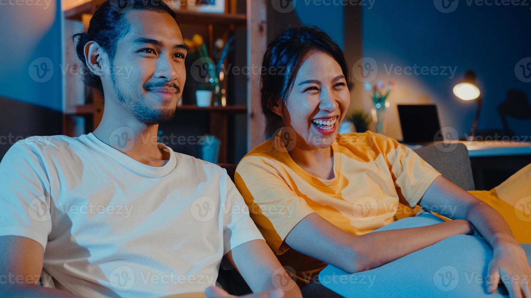 romântico asiático casal homem e mulher sorrir e rir deitar no sofá na sala de estar à noite assistir filme de comédia na televisão juntos em casa. casal casado estilo de vida familiar, conceito de ficar em casa. foto