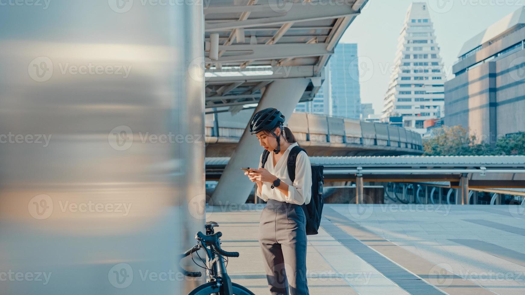 sorriso asiática empresária com mochila usar telefone inteligente olhar câmera na cidade ficar na rua com bicicleta ir trabalhar no escritório. garota esporte usa o telefone para o trabalho. comutar para trabalhar, viajante de negócios na cidade. foto