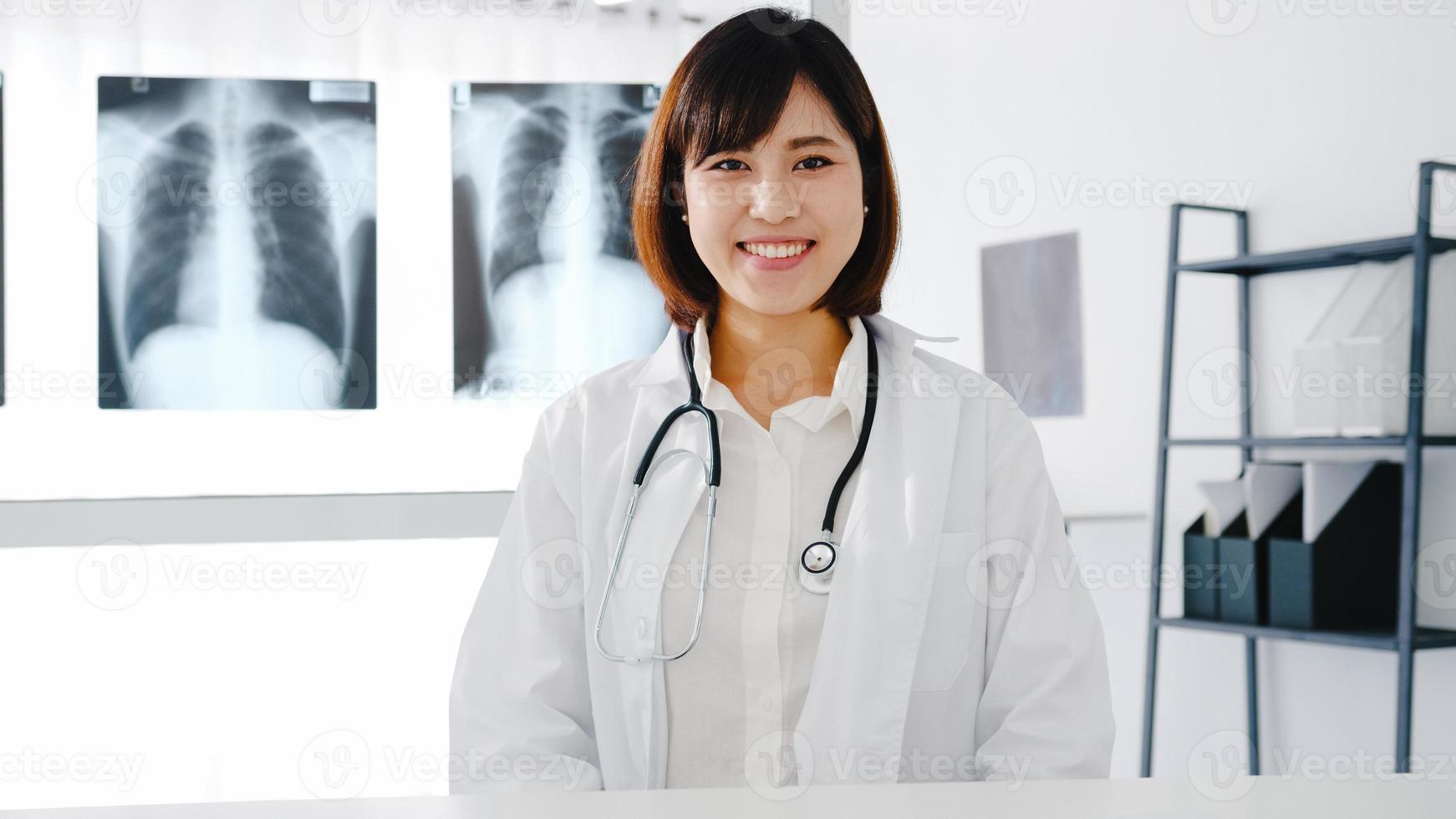 confiante jovem médica asiática em uniforme médico branco com estetoscópio, olhando para a câmera e sorrindo durante a videoconferência com o paciente no hospital de saúde. consultoria e conceito de terapia. foto