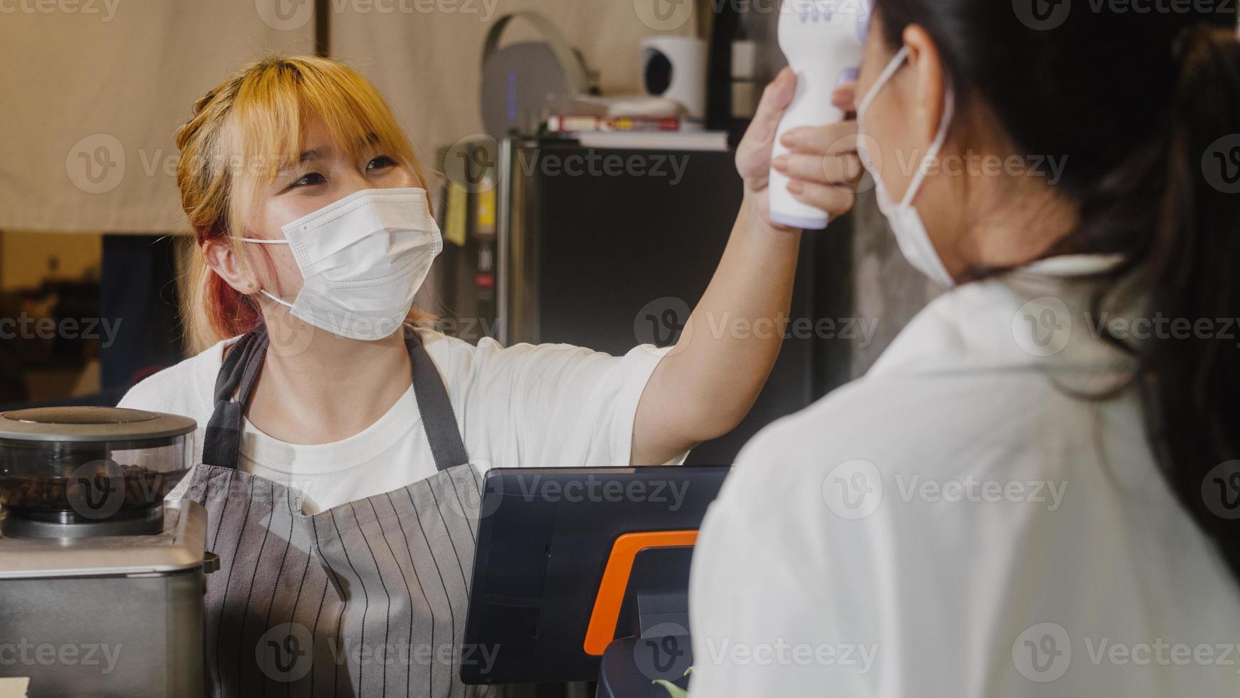 jovem funcionária do restaurante asiático usando máscara protetora usando o verificador de termômetro infravermelho ou pistola de temperatura na testa do cliente antes de entrar. estilo de vida novo normal após o vírus corona. foto