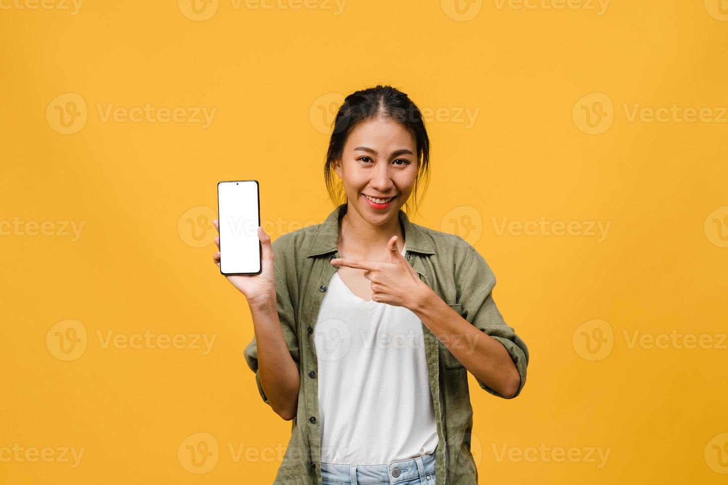 jovem senhora asiática mostra tela vazia do smartphone com expressão positiva, sorri amplamente, vestida com roupas casuais, sentindo felicidade sobre fundo amarelo. telefone celular com tela branca na mão feminina. foto