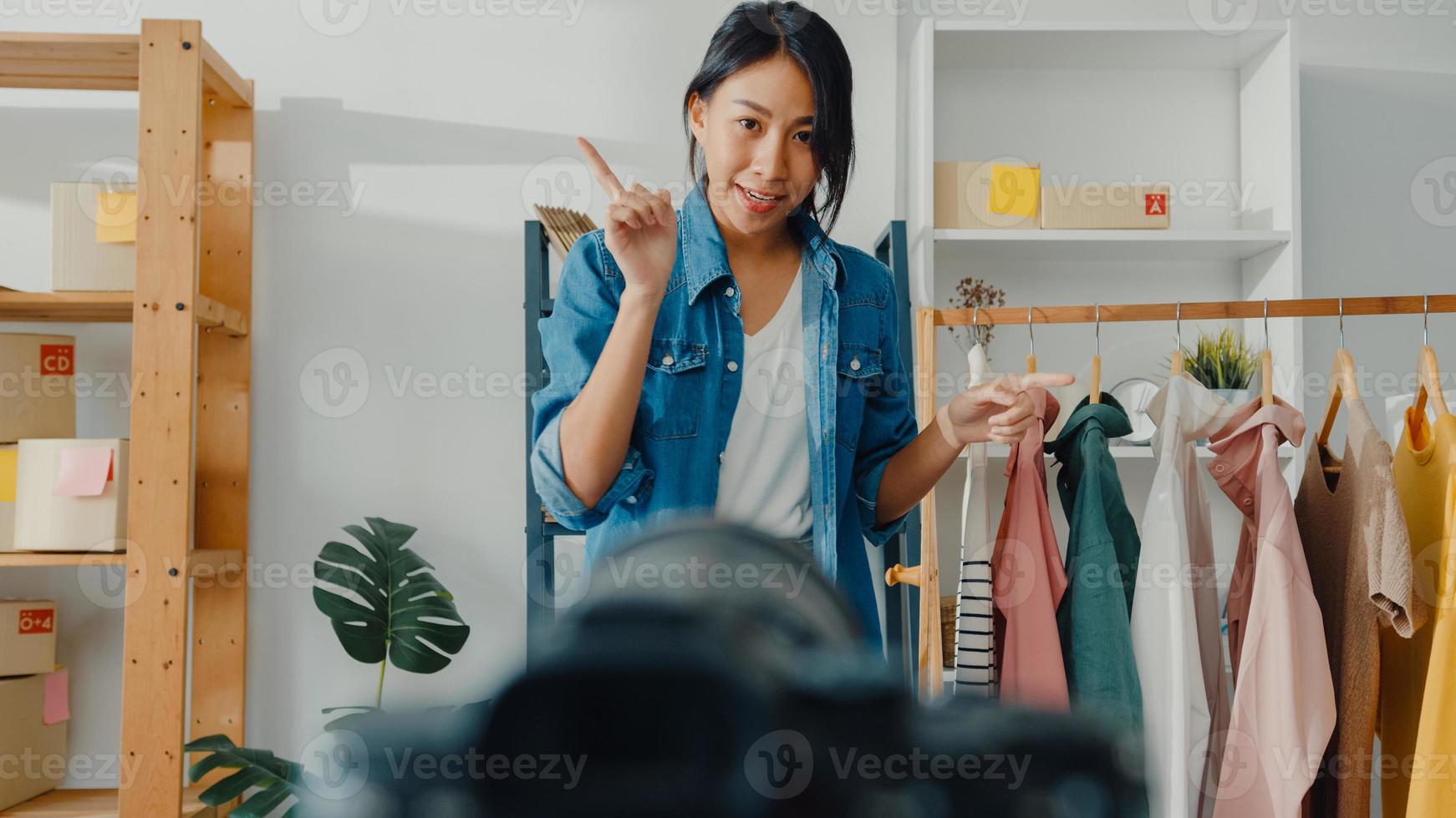 jovem estilista da Ásia usando o telefone celular, recebendo o pedido de compra e mostrando as roupas, gravando vídeo com transmissão ao vivo online com a câmera. proprietário de uma pequena empresa, conceito de entrega de mercado online. foto