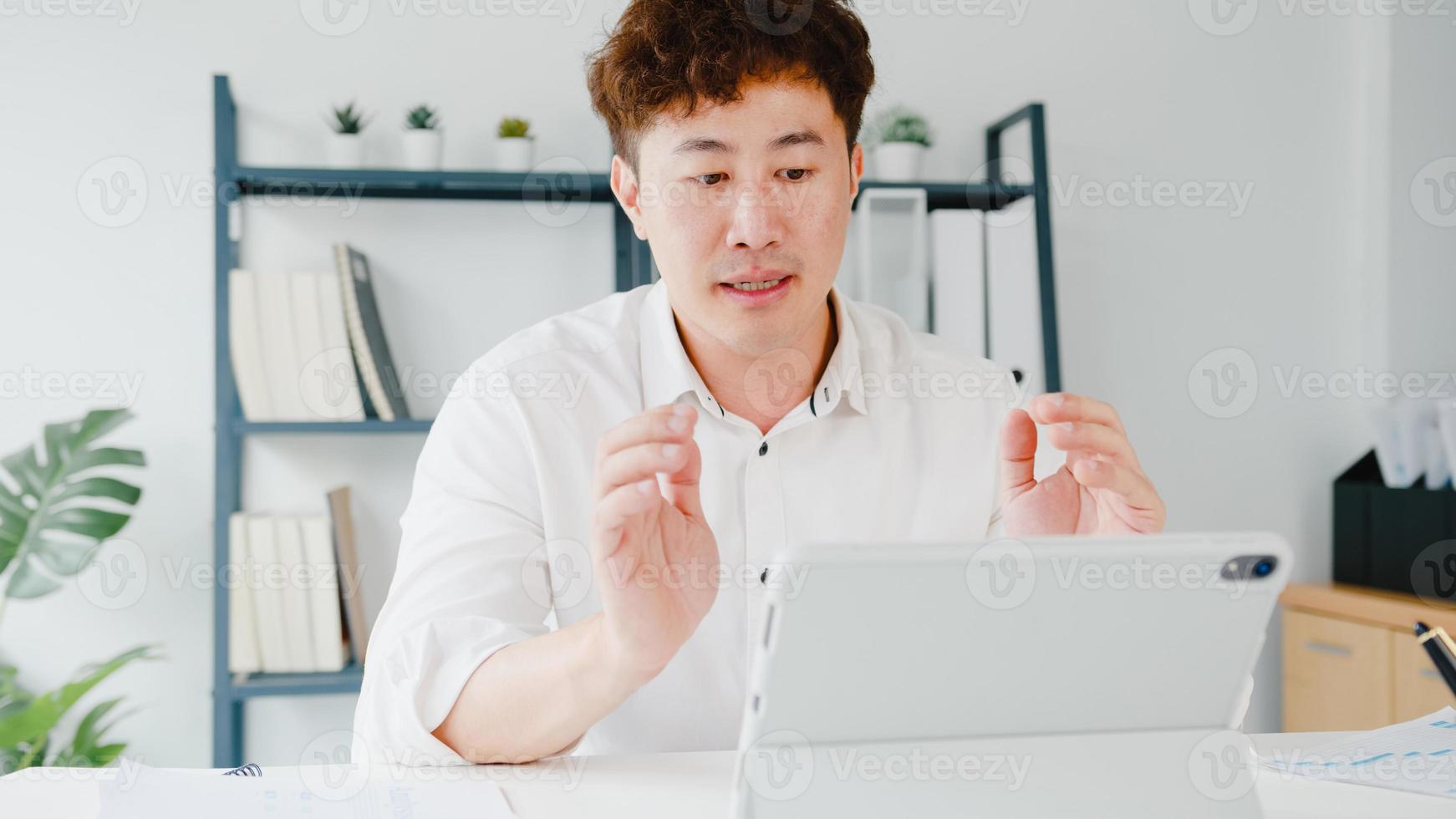 jovem empresário asiático usando tablet conversa com colegas sobre o plano de videochamada enquanto inteligente trabalhando em casa na sala de estar. auto-isolamento, distanciamento social, quarentena para prevenção do vírus corona. foto