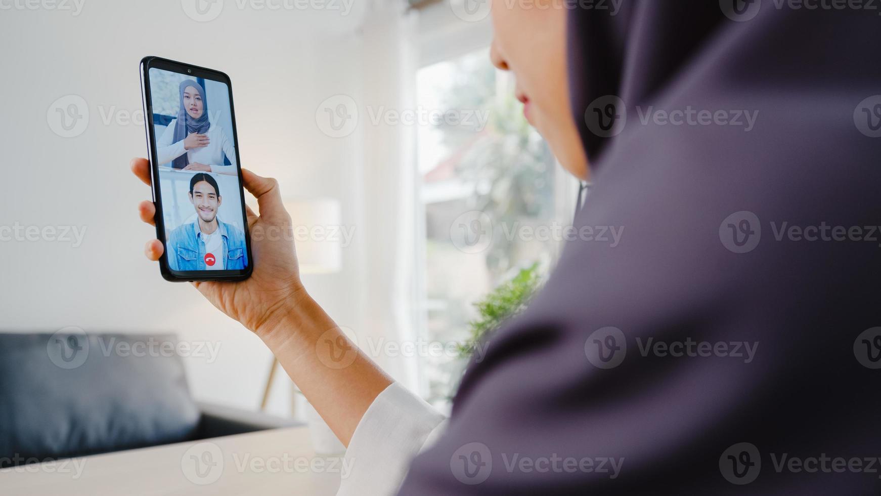 jovem empresária muçulmana asiática usando telefone inteligente, conversa com um colega por videochat para uma reunião on-line enquanto trabalha remotamente em casa na sala de estar. distanciamento social, quarentena para o vírus corona. foto