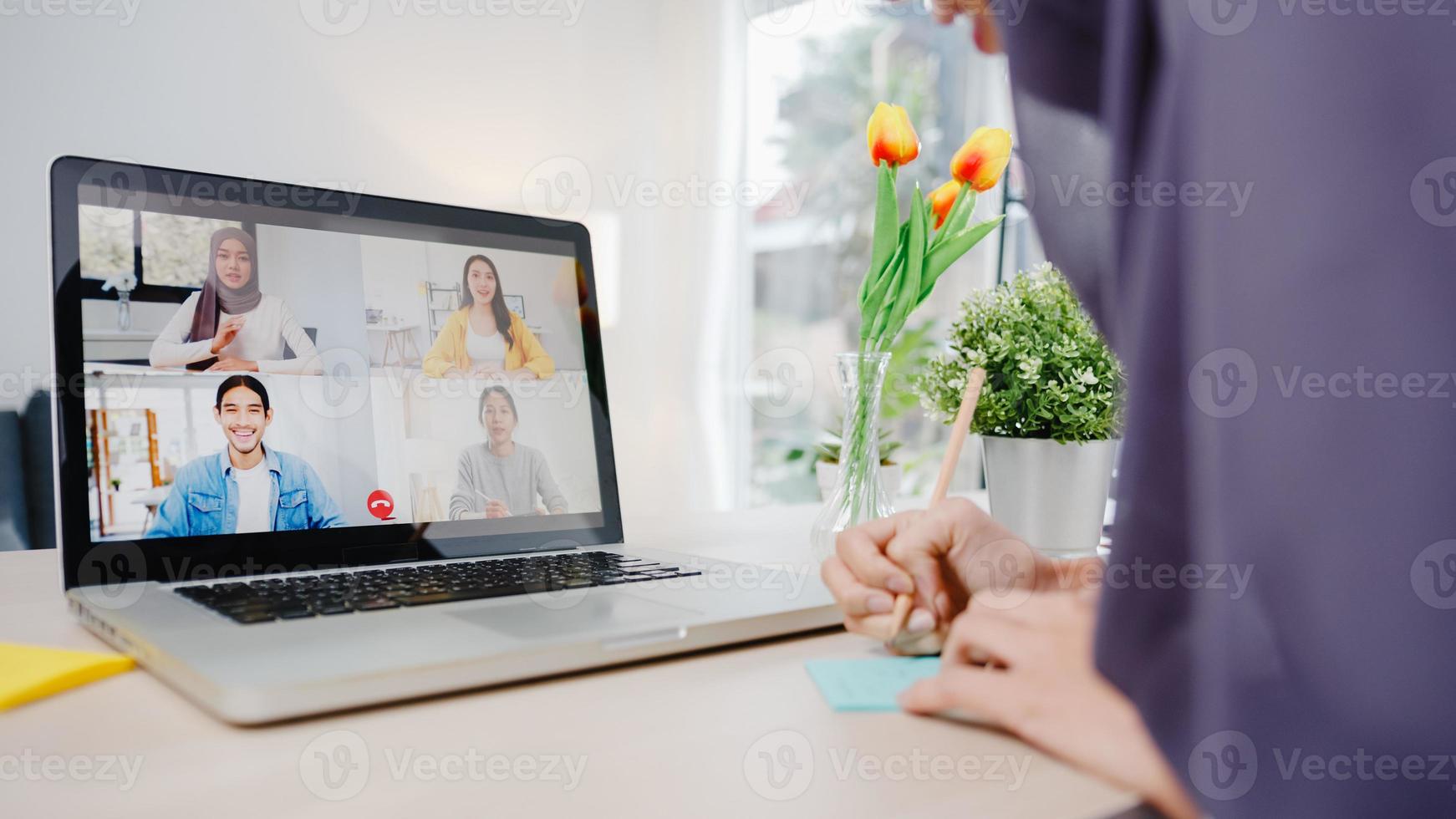 empresária muçulmana asiática usando laptop fala com um colega sobre plano por videochamada, brainstorm reunião on-line enquanto trabalha remotamente de casa na sala de estar. distanciamento social, quarentena para o vírus corona foto