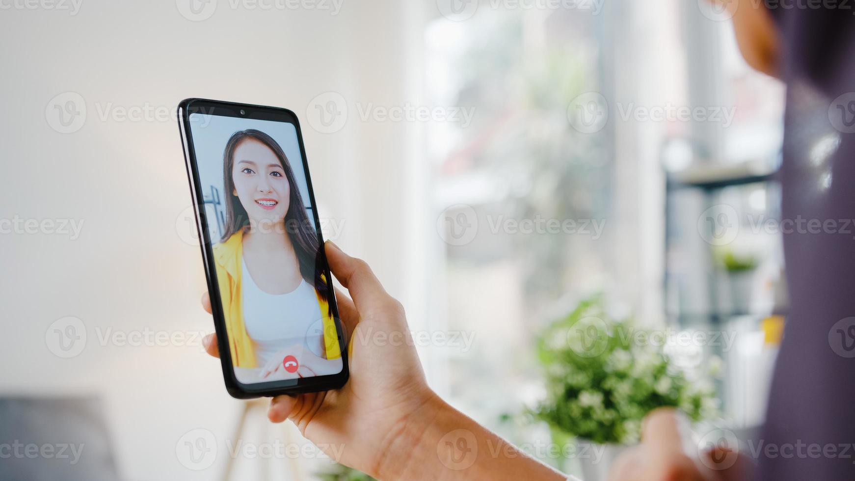 jovem empresária muçulmana asiática usando telefone inteligente, conversa com um amigo por videochat, brainstorm, reunião on-line, enquanto trabalha remotamente em casa na sala de estar. distanciamento social, quarentena para o vírus corona. foto