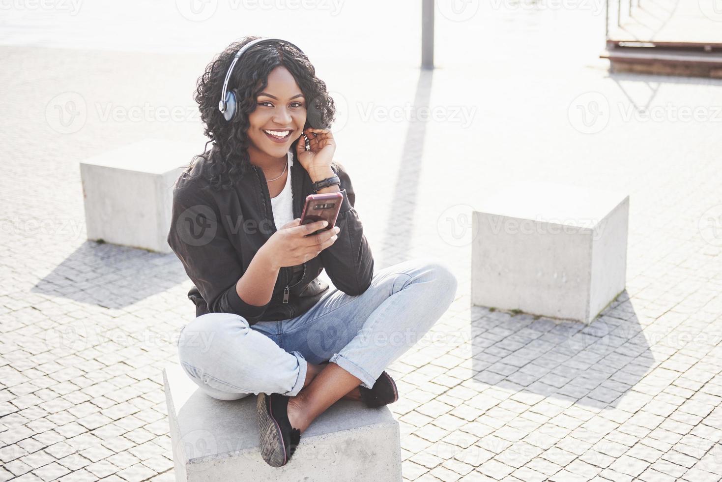 retrato de uma bela jovem afro-americana sentada na praia ou lago e ouvindo música em seus fones de ouvido foto