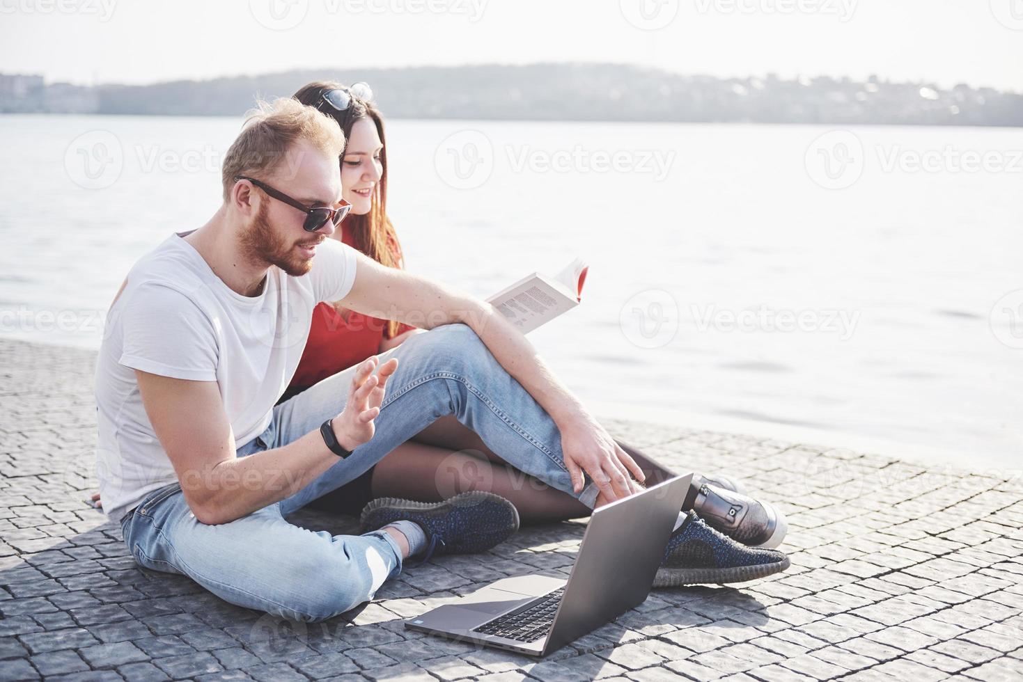 dois alunos, rapaz e moça, sentados ao ar livre, desfrutando de um laptop, estudando ao ar livre em um dia ensolarado foto