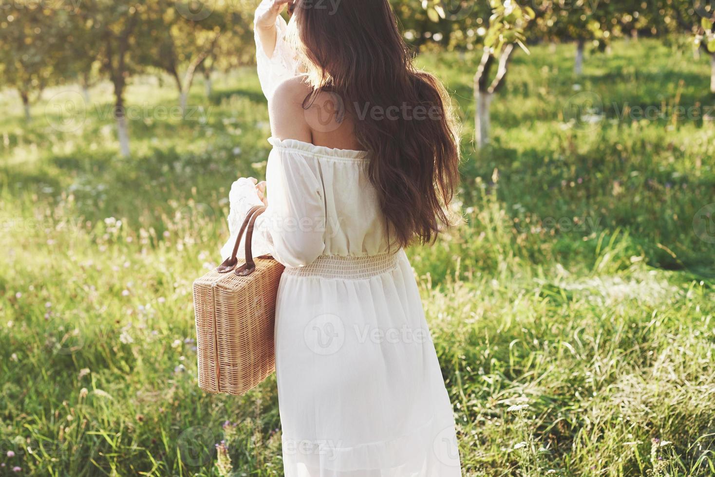 uma jovem com um vestido longo branco está caminhando no jardim. lindo pôr do sol através das folhas das árvores foto